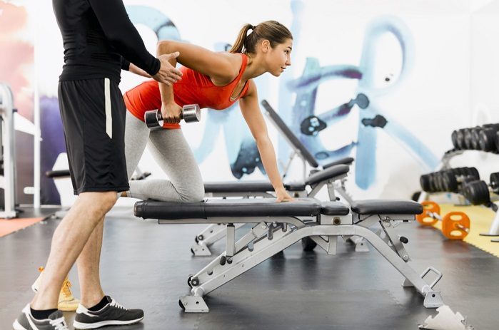 woman performing dumbbell rows with personal trainer in her local gym