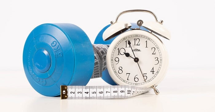 time for exercises blue dumbbell, measuring tape and an alarm clock on a white surface
